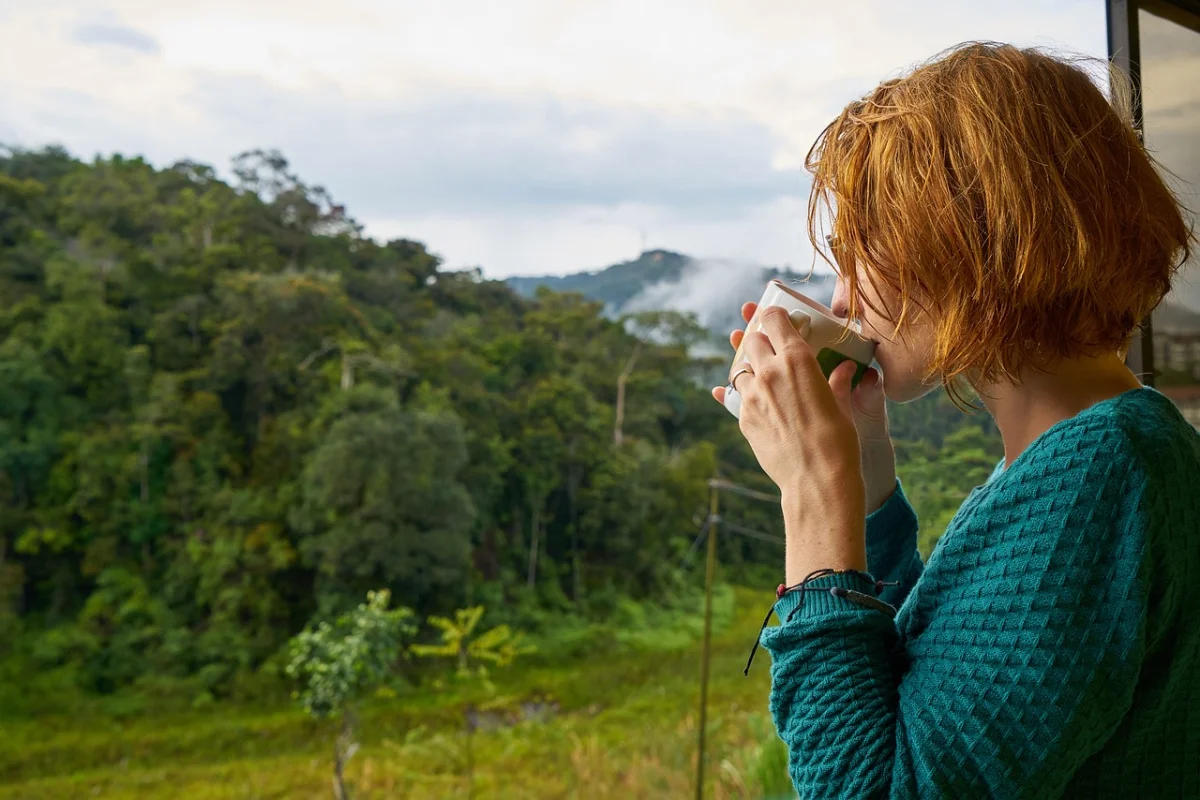 A woman in a teal sweater sipping tea, gazing out at a lush green landscape.
