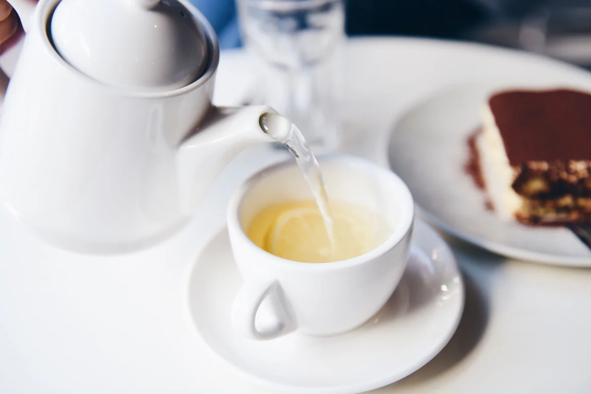 Close-up of tea being poured into a white cup from a teapot, with a slice of cake on a plate in the background.