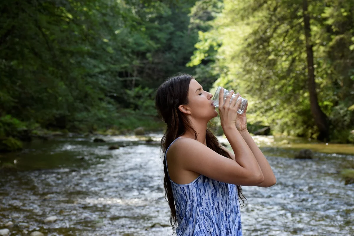 A woman drinking from a clear glass in a natural, forested riverside setting.