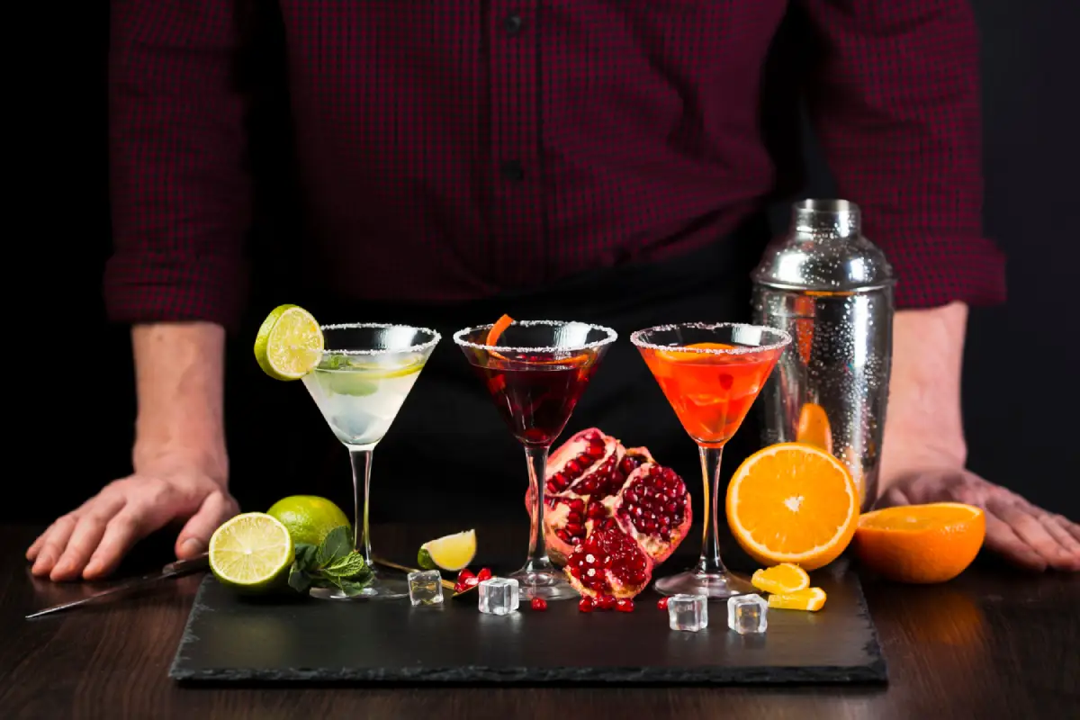 An array of colorful cocktails with garnishes on a dark slate, with a bartender in the background