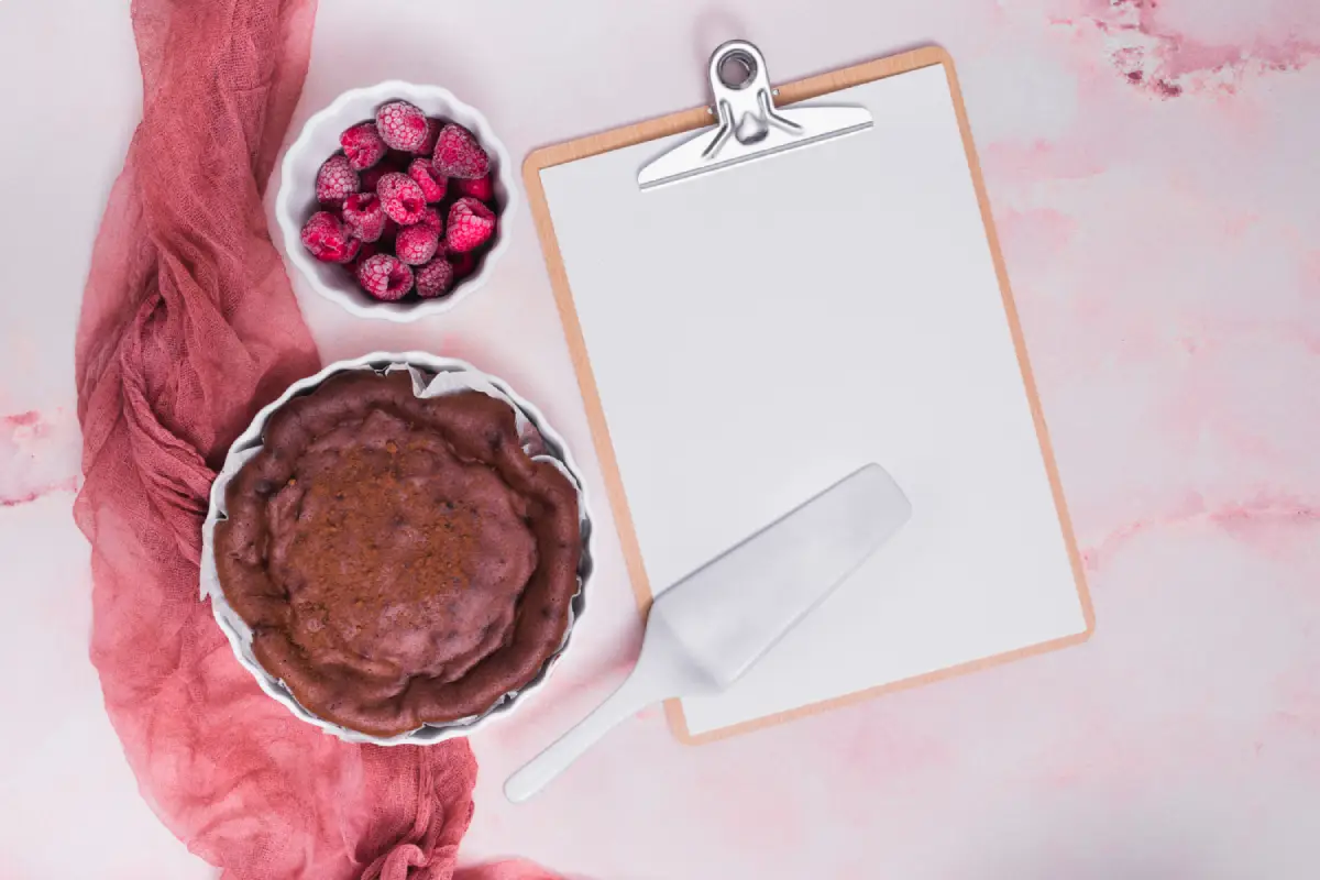 Baked cake in a pie dish alongside a bowl of raspberries and a spatula, with a clipboard and blank paper on a pink textured background, ready for noting nutritional information.
