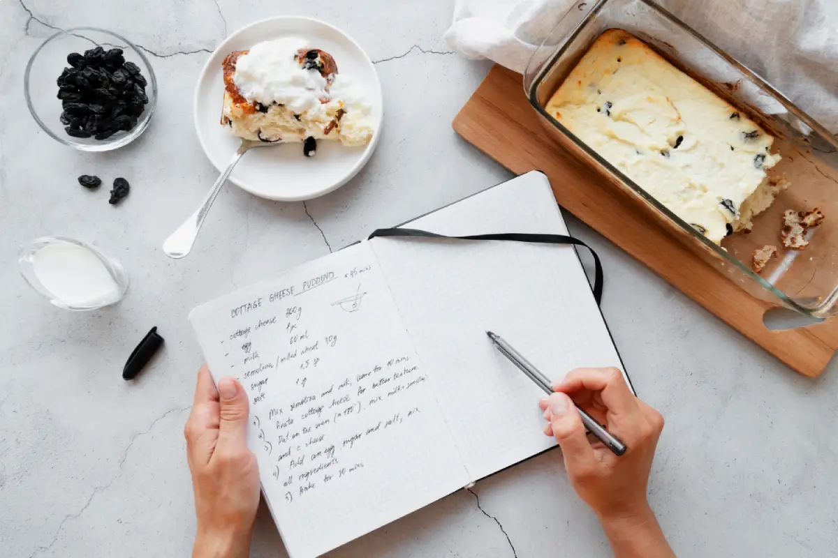Hands writing in a recipe book with notes on making cottage cheese pudding, alongside a bowl of raisins, a dish of pudding, and a glass of milk on a marble surface.