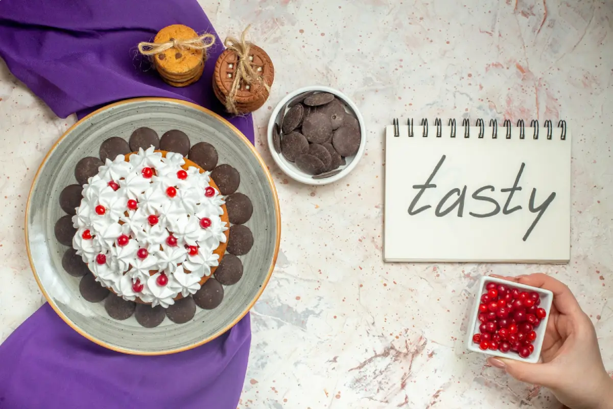 Hand adding red currants to a beautifully decorated cake with whipped cream, surrounded by cookies, chocolates, and a 'tasty' note, depicting a personal touch in dessert presentation.
