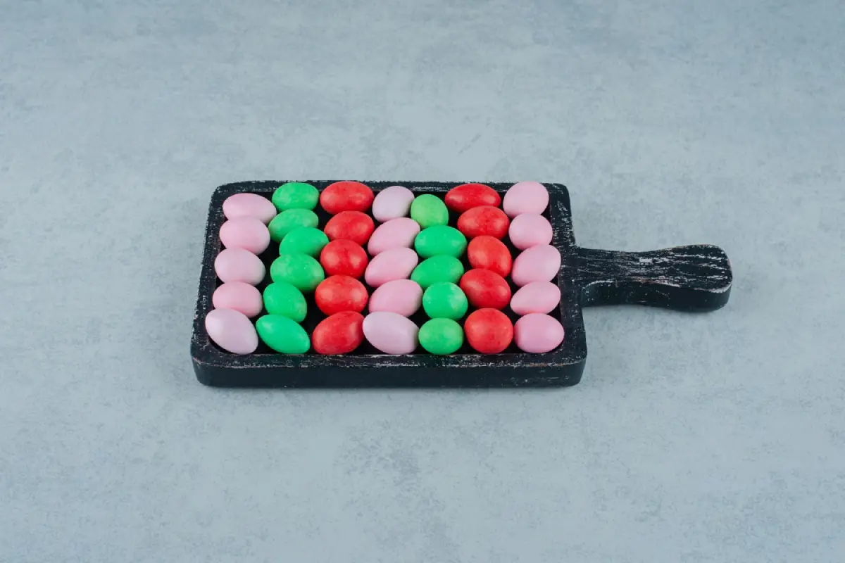 A neatly arranged pattern of pink, green, and red round candies on a dark wooden cutting board against a light textured background.