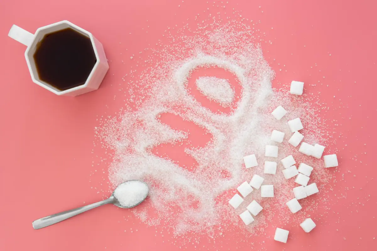 The word "NO" spelled out in granulated sugar, with a spoonful of sugar and cubes on a pink background, next to a cup of coffee.