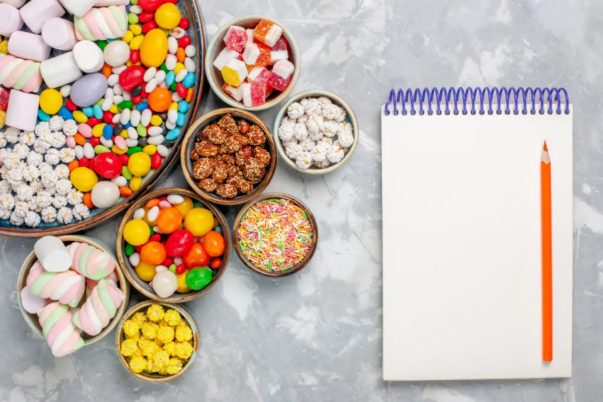 An assortment of colorful candies and sweets laid out next to an open notebook and pencil on a light marbled surface.