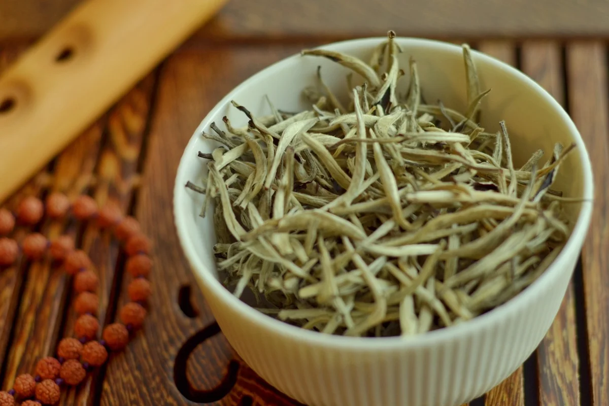 A bowl full of dried white tea leaves on a wooden table, accompanied by prayer beads.