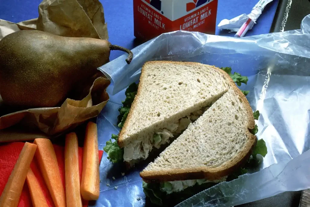 Panera Tuna Salad Recipe-inspired lunch featuring a whole grain bread sandwich with tuna salad, alongside fresh carrot sticks, a ripe pear, and a carton of low-fat milk.