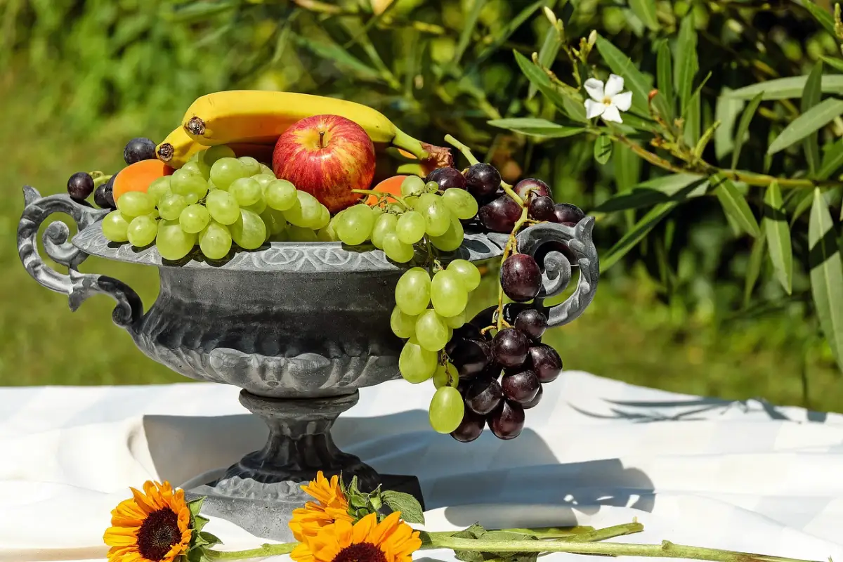 Ornate black fruit bowl filled with bananas, grapes, and an apple, adorned with sunflowers on a table. Image by Ilo from Pixabay