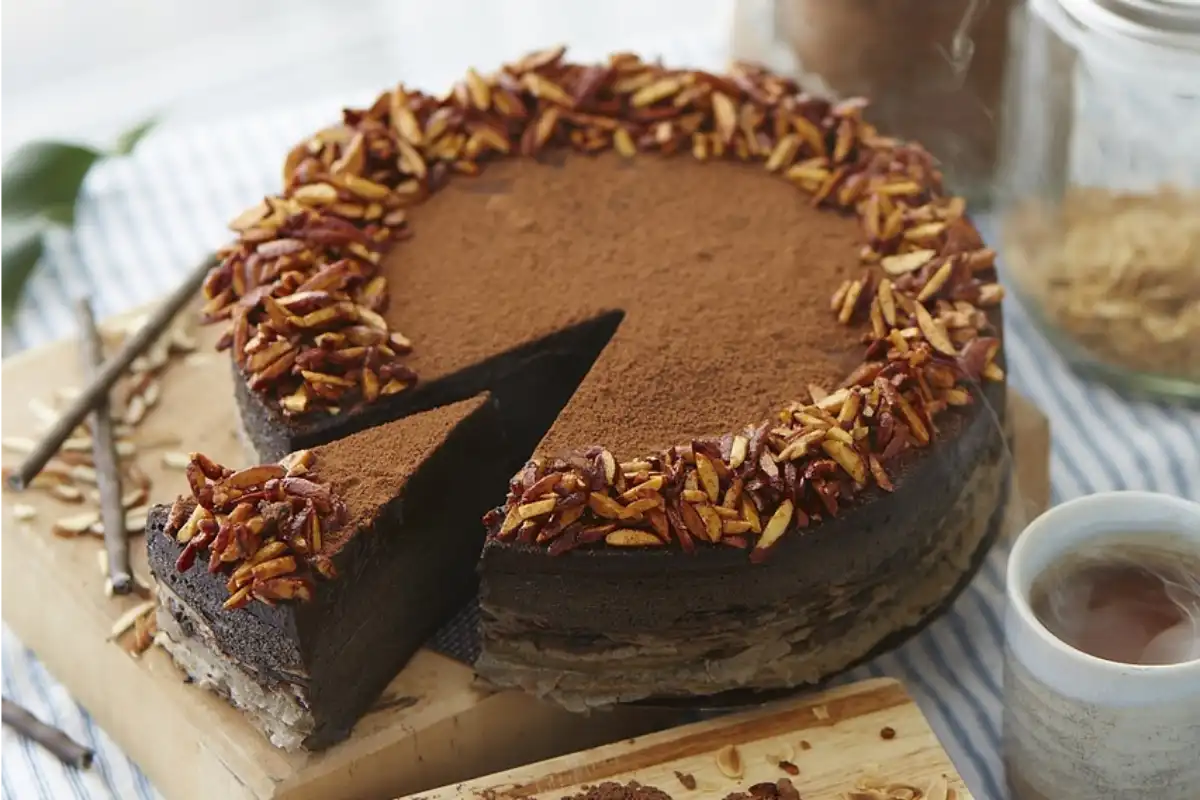 Elegant candy cake topped with a dusting of cocoa powder and a ring of chopped pecans on a wooden board with a slice being served.
