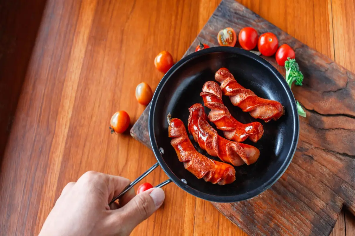 Chef's hands cooking Red Hots sausages in a skillet with tomatoes on the side.
