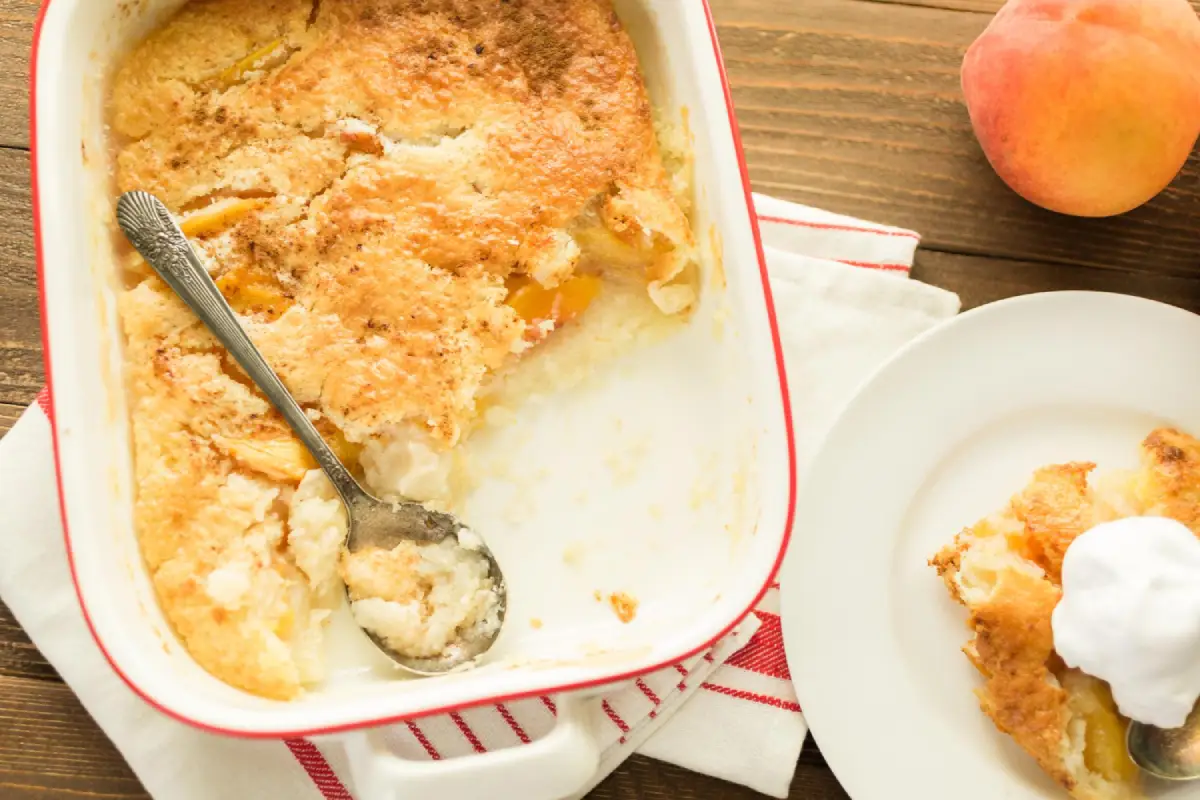 Freshly baked peach cobbler in a dish with a serving spoon and whipped cream on the side.