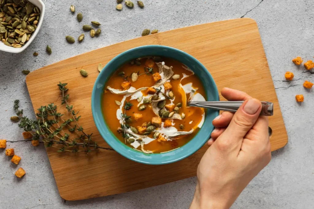 A bowl of vegetable soup garnished with herbs and seeds, a hand holding a spoon