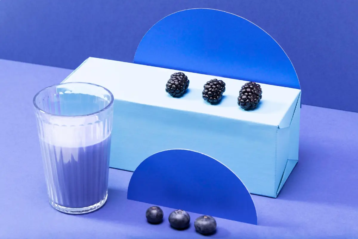 A geometrically arranged still life of blackberries, blueberries, and a glass of smoothie on a dual-tone blue background.