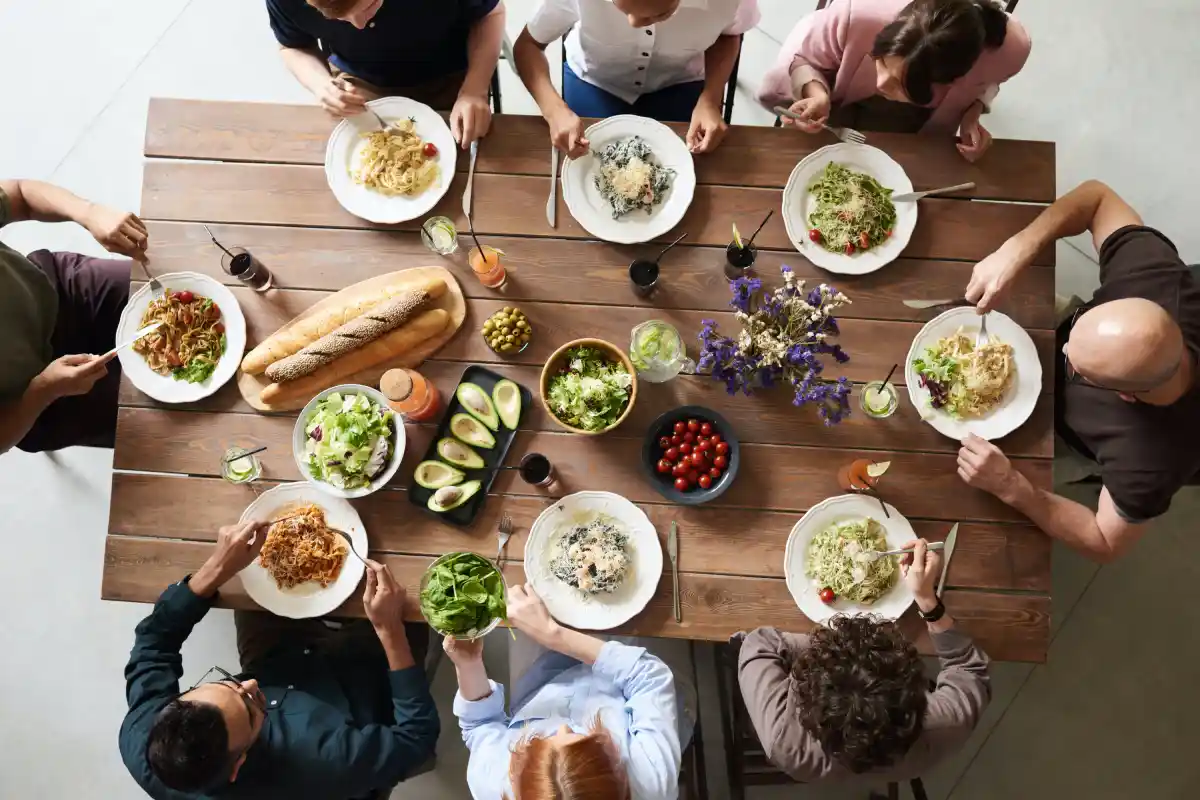 Diverse group of people sharing a meal with various pasta dishes around a wooden table.