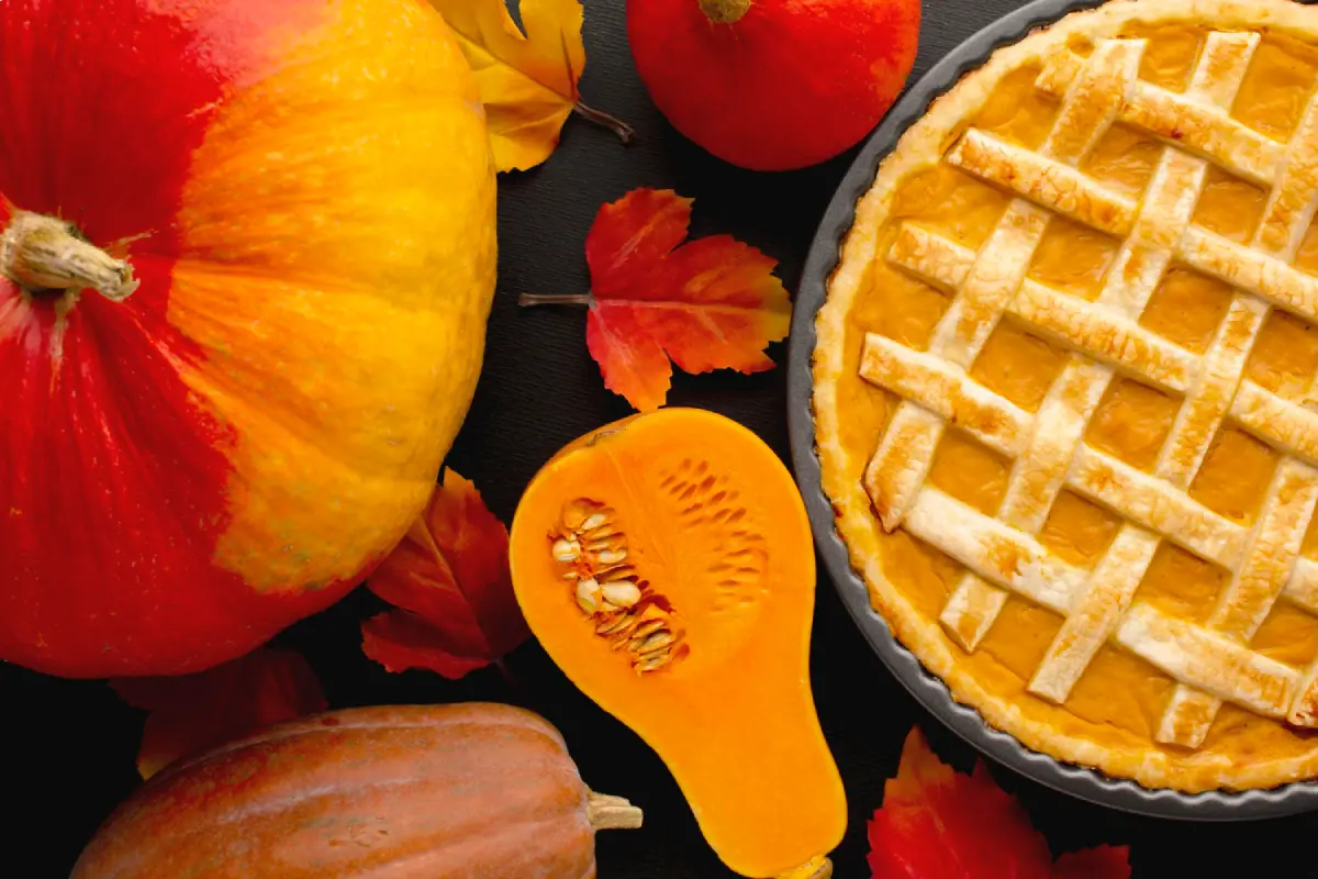 A freshly baked pumpkin pie beside whole and sliced pumpkins on a dark background with autumn leaves