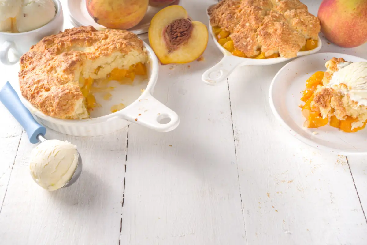Half-eaten peach cobbler in a white baking dish beside fresh peaches and a scoop of ice cream.
