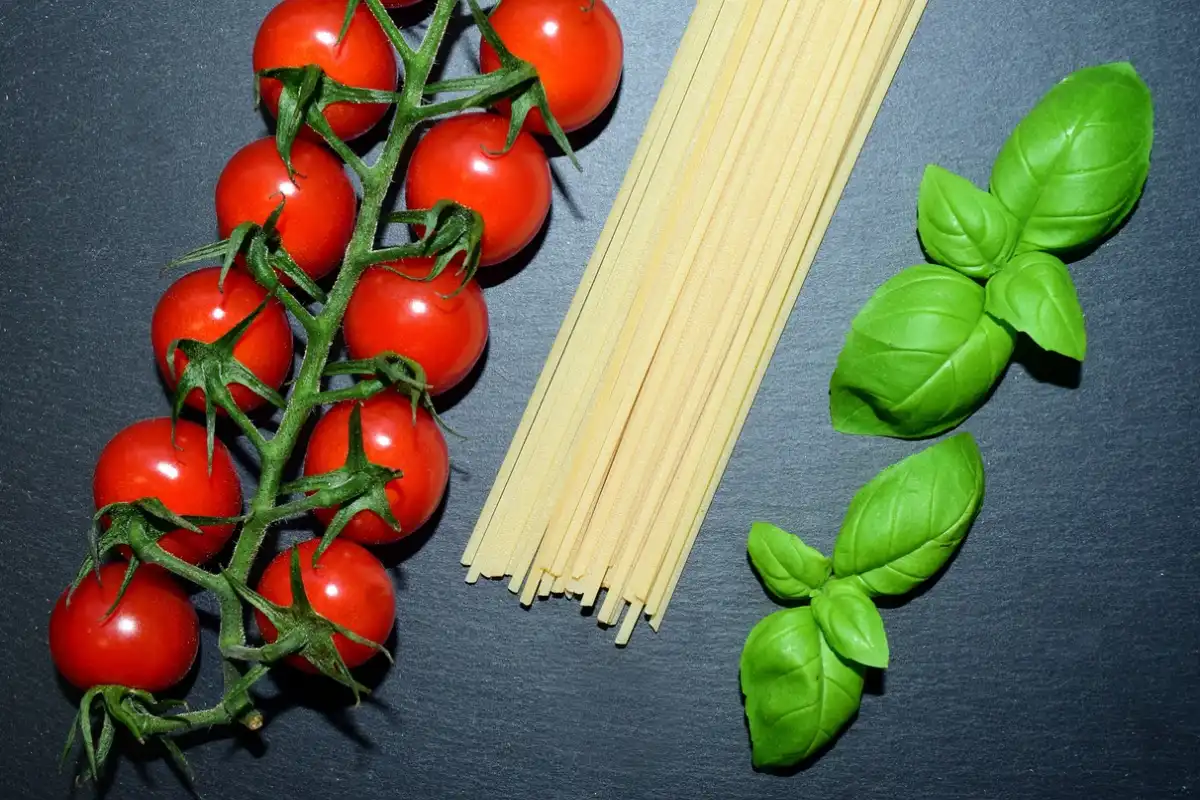A raw setup of spaghetti, fresh basil, and ripe cherry tomatoes on a slate background, representing Italian culinary staples.