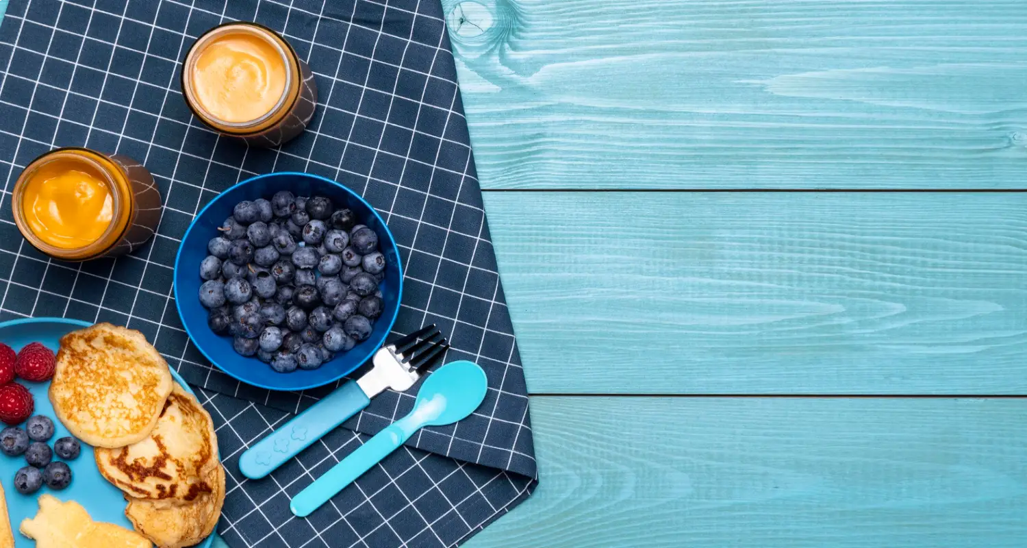 A top-down view of a breakfast setup with blueberries, pancakes, and jars of baby food on a checkered cloth and teal wooden surface