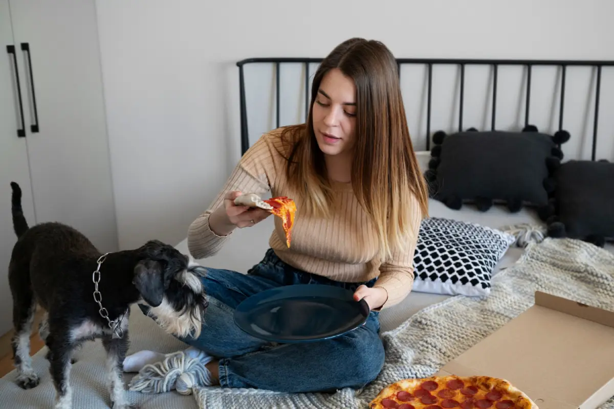 Woman on bed with pizza, dog looking on, highlighting foods toxic to dogs.