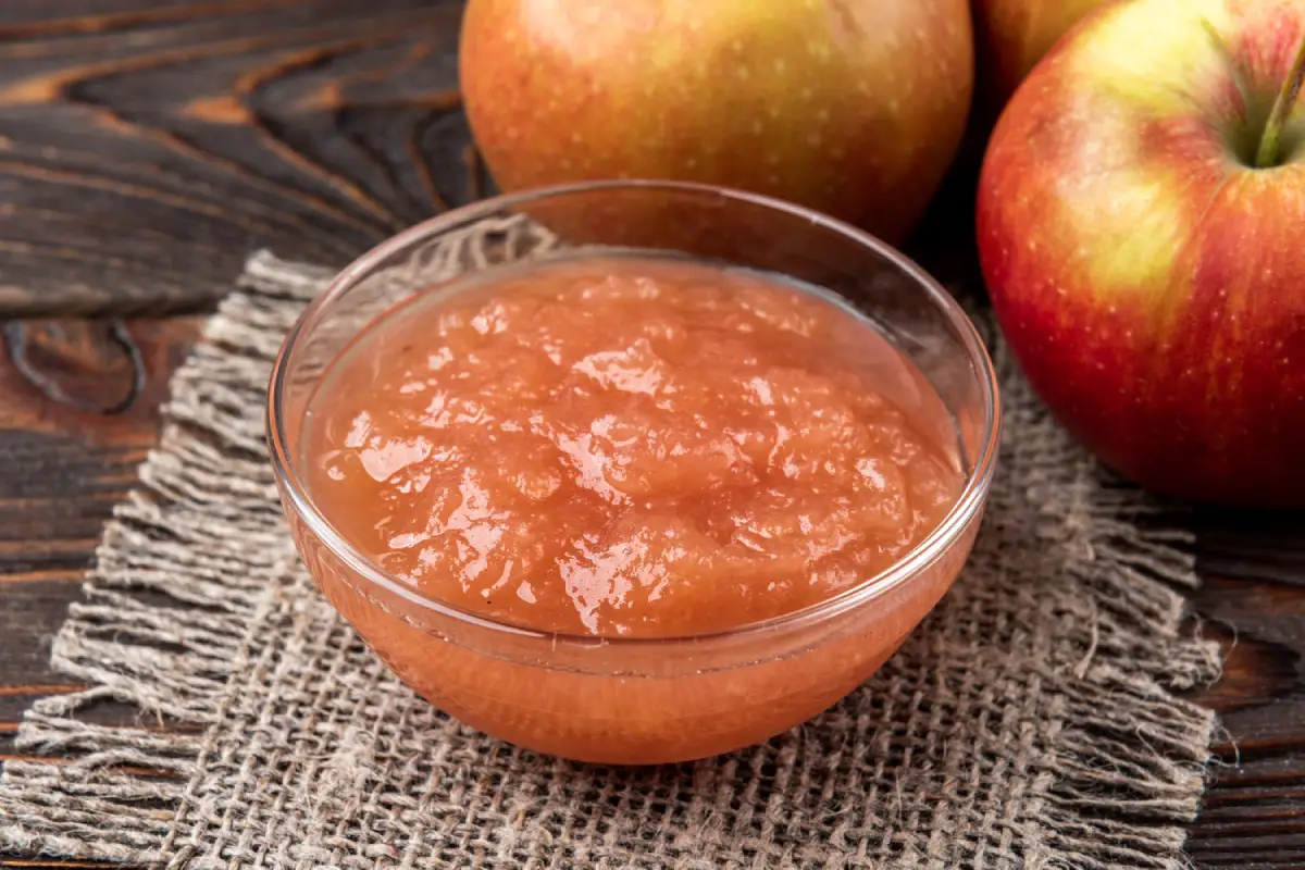 A clear bowl of homemade apple sauce with whole apples on a dark wooden surface.