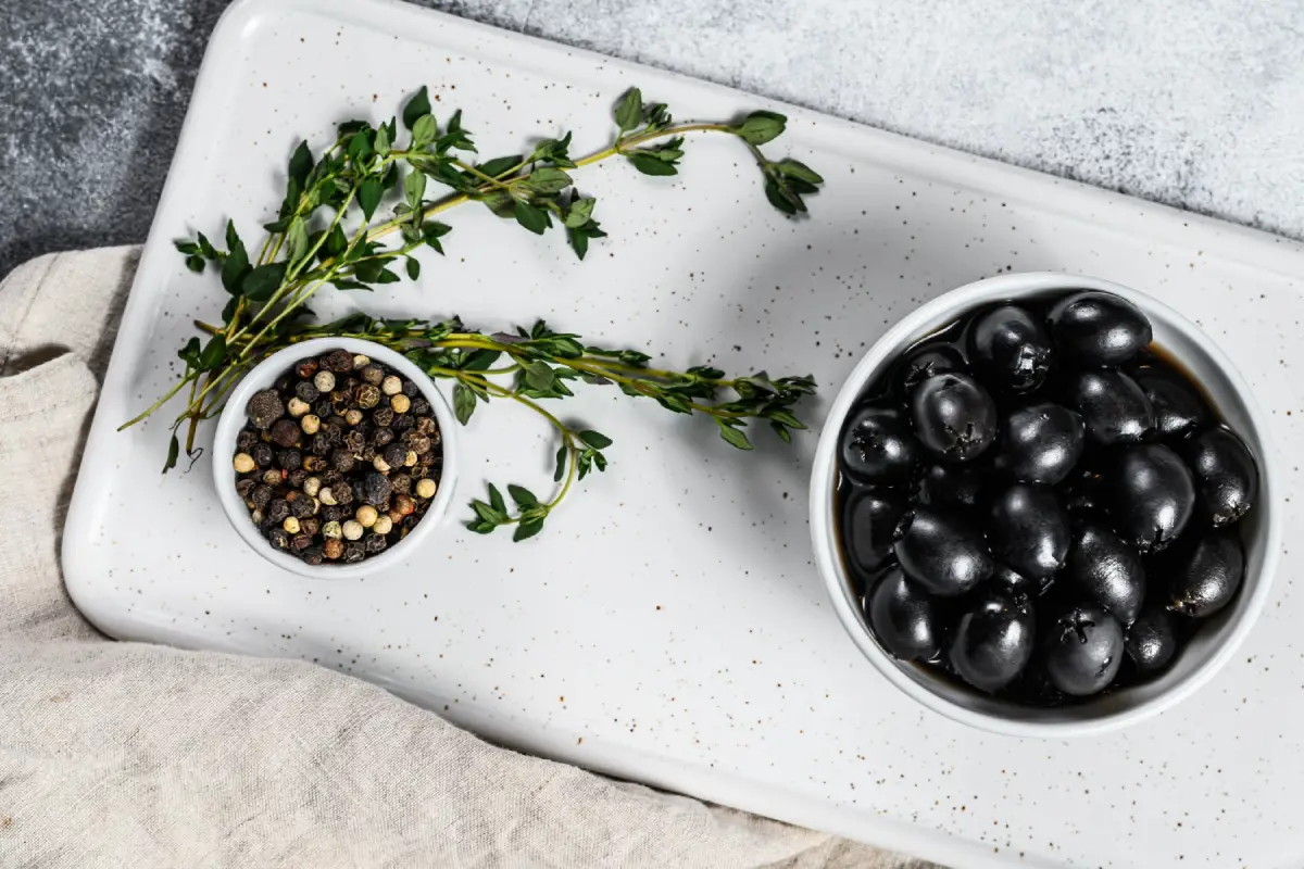 A bowl of black olives next to mixed peppercorns and fresh thyme on a white chopping board