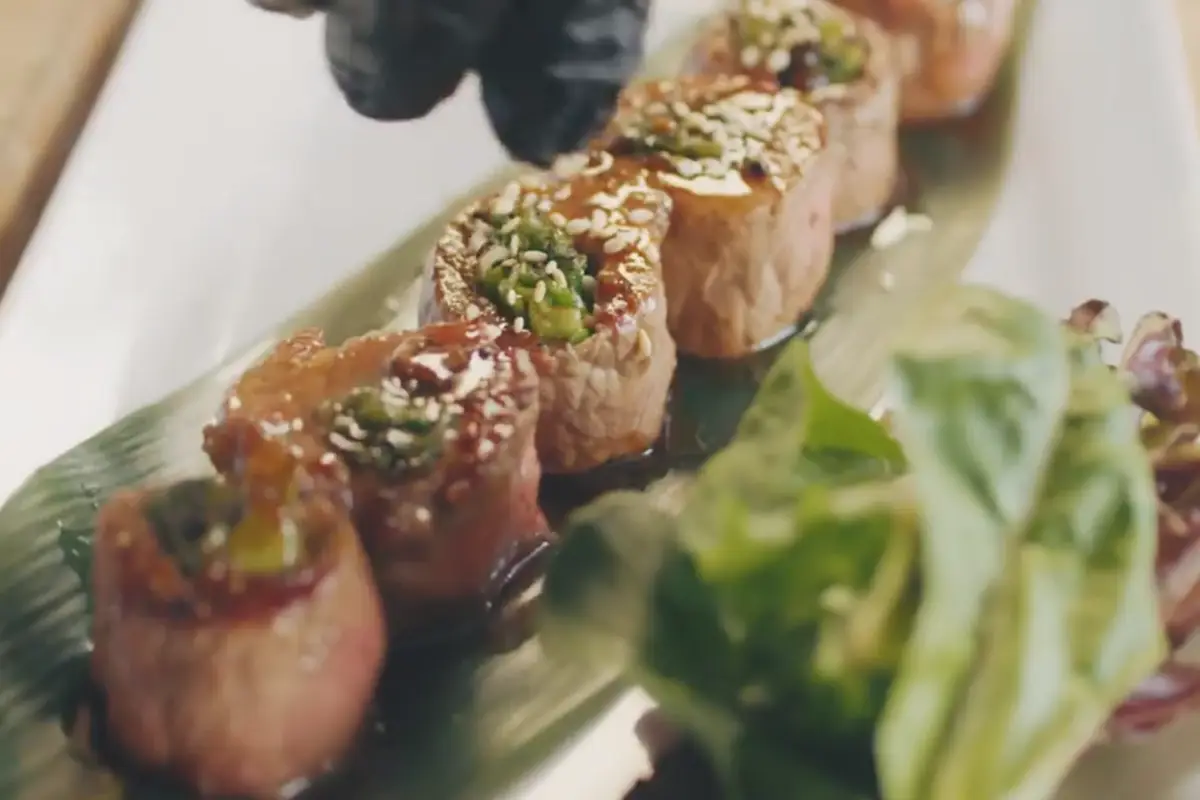 Slices of beef negimaki garnished with sesame seeds and green onions, served on a bamboo leaf.