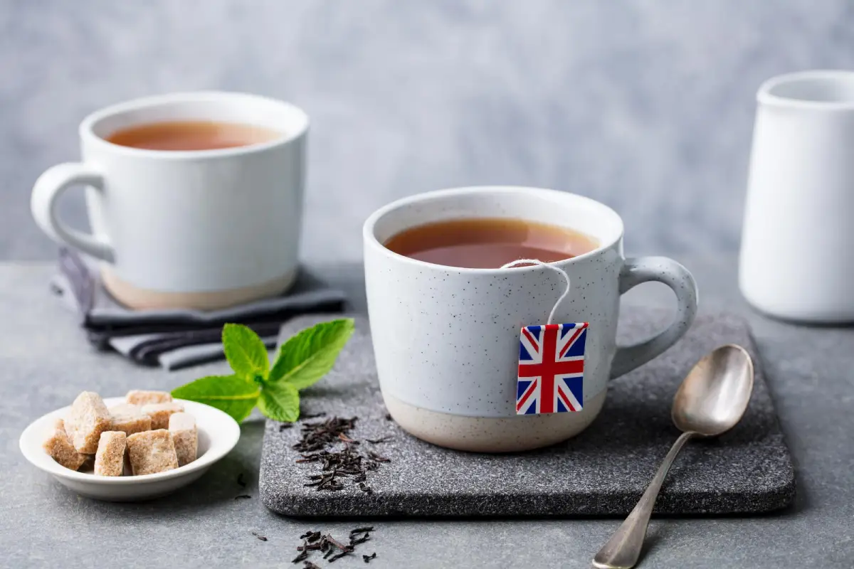 Two mugs of Earl Grey tea with a British flag tea tag on a grey countertop with loose tea leaves and brown sugar cubes.