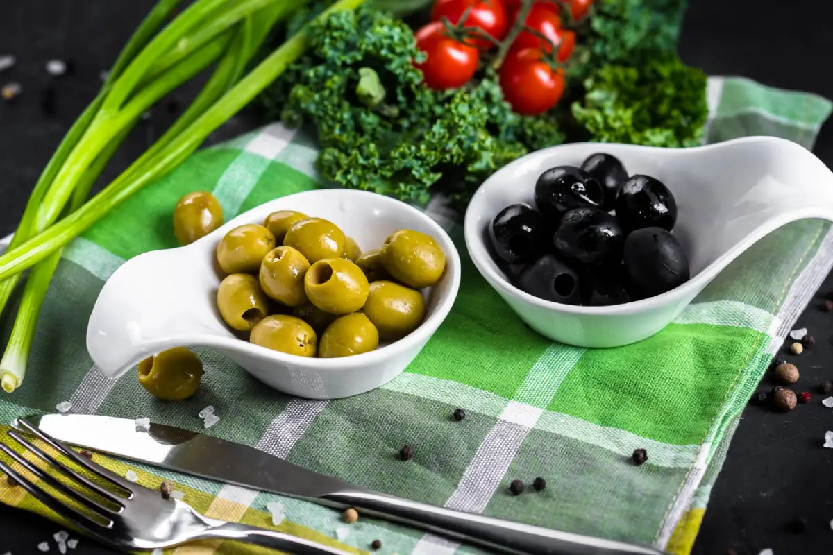 A comparison of black and green olives in white bowls, with fresh vegetables and cutlery on a checkered cloth.