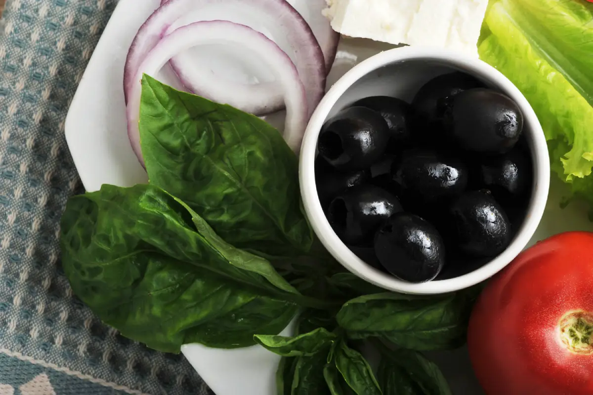 Black olives in a bowl accompanied by fresh basil, onions, and tomato, ingredients for a healthy meal.