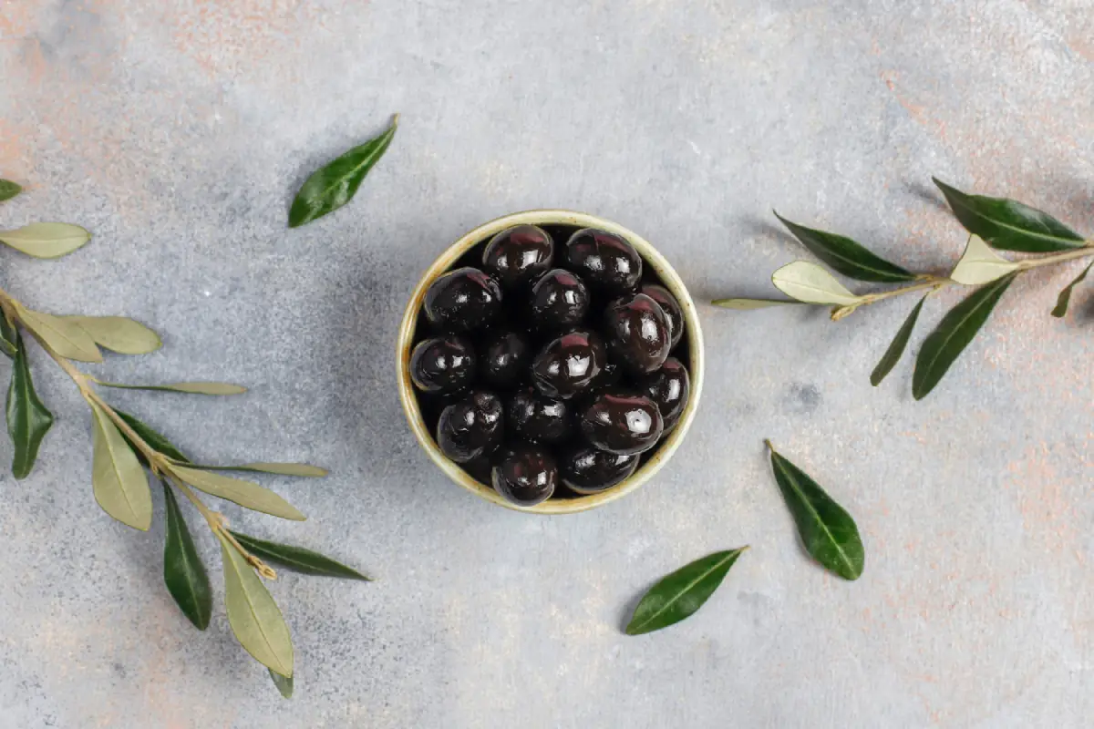 Bowl of black olives surrounded by olive leaves on a textured surface.