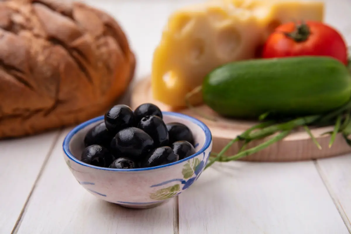 Bowl of black olives in focus with cheese, tomato, cucumber, and bread in the background