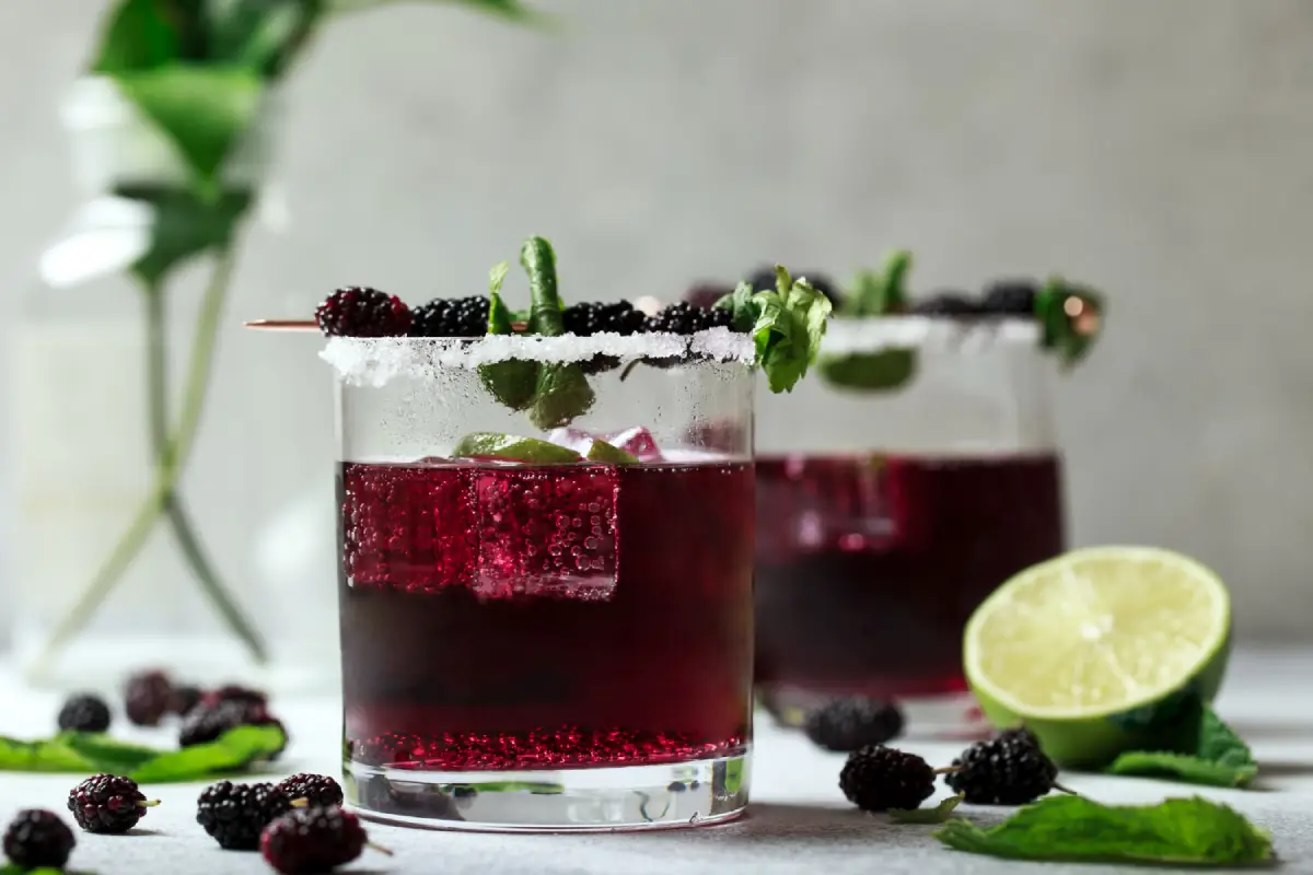 An elegant blackberry moonshine cocktail garnished with fresh berries and mint, served with a rim of sugar on a light background.