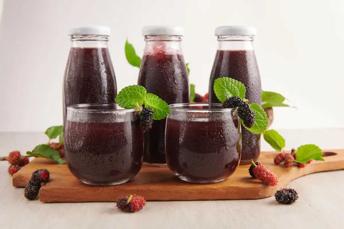 Bottles and glasses of blackberry moonshine with mint leaves and berries on a wooden serving tray.