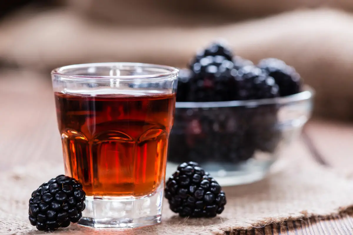 Homemade blackberry moonshine in a shot glass beside fresh blackberries on a rustic wooden table.