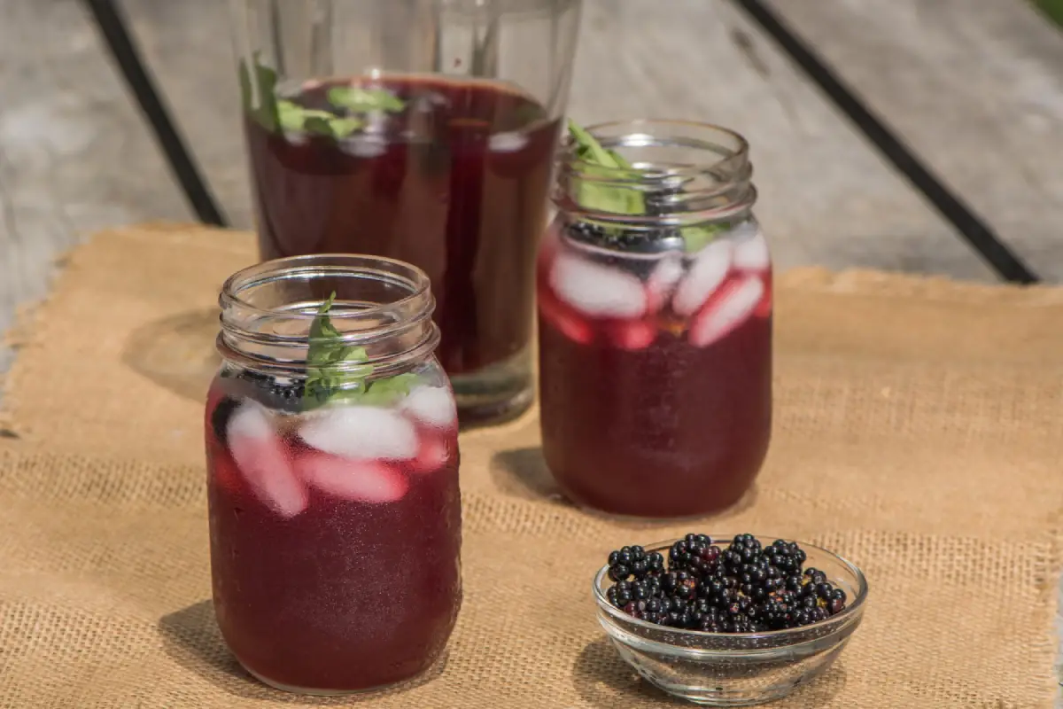 Refreshing blackberry moonshine cocktails with ice and mint in mason jars, accompanied by a bowl of fresh blackberries on a burlap cloth.