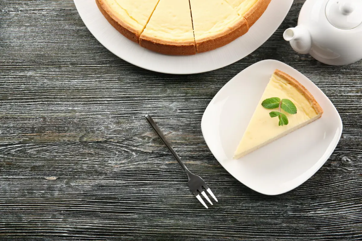 A slice of classic vanilla cheesecake on a rustic wooden table with a full cake in the background.