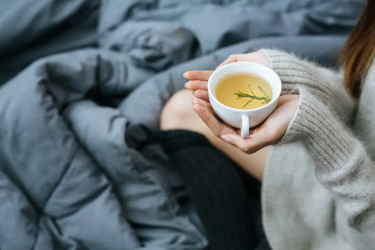 Person holding a cup of tea with a sprig of rosemary, cozy in bed with gray linens.