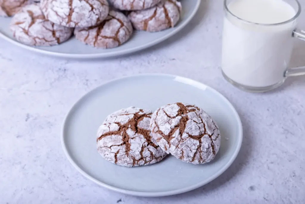 Chocolate cool whip cookies dusted with powdered sugar on a plate, served with a mug of milk.