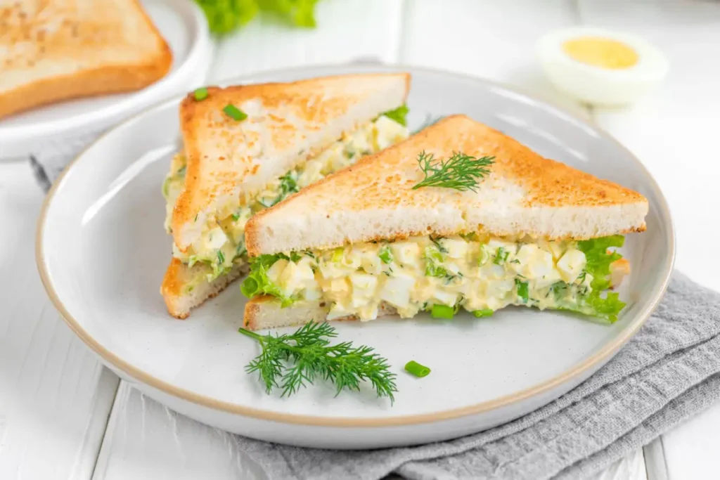 Egg salad sandwich with toasted bread and fresh lettuce on a white wooden background.