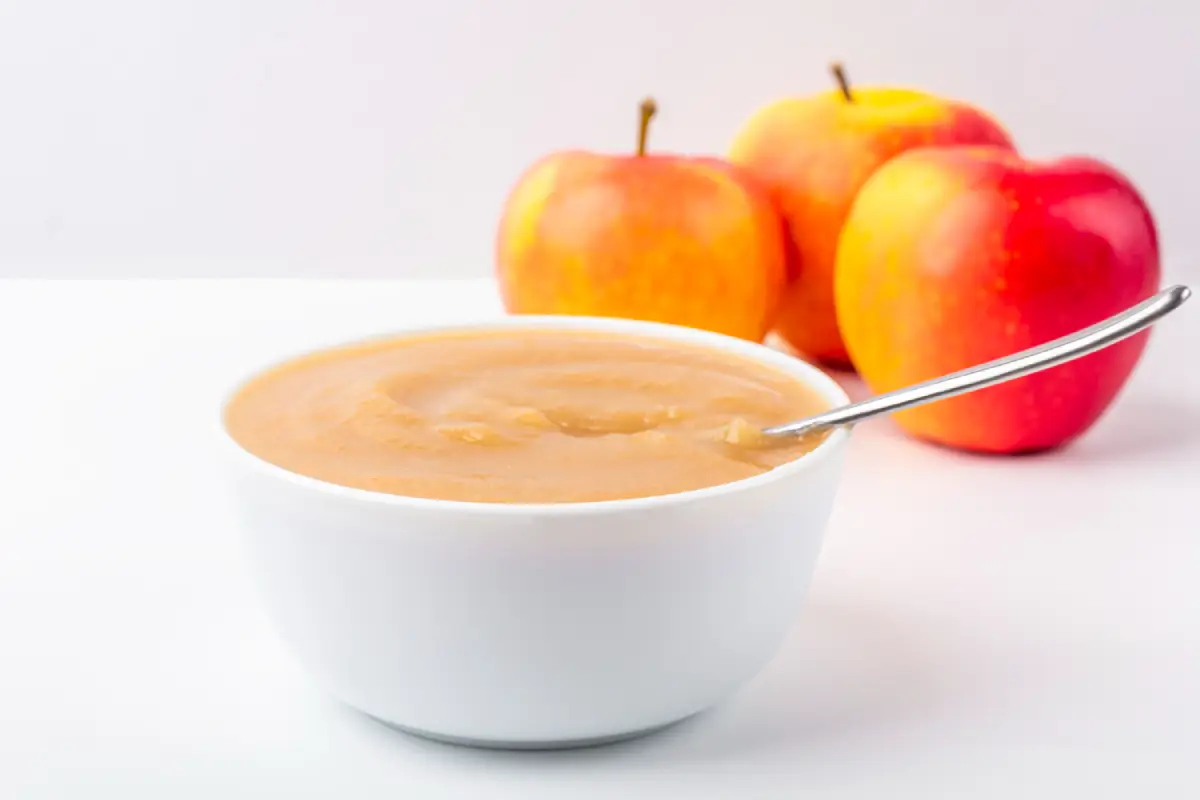 Creamy homemade apple sauce in a white bowl with a spoon, with fresh apples in the background on a white surface.