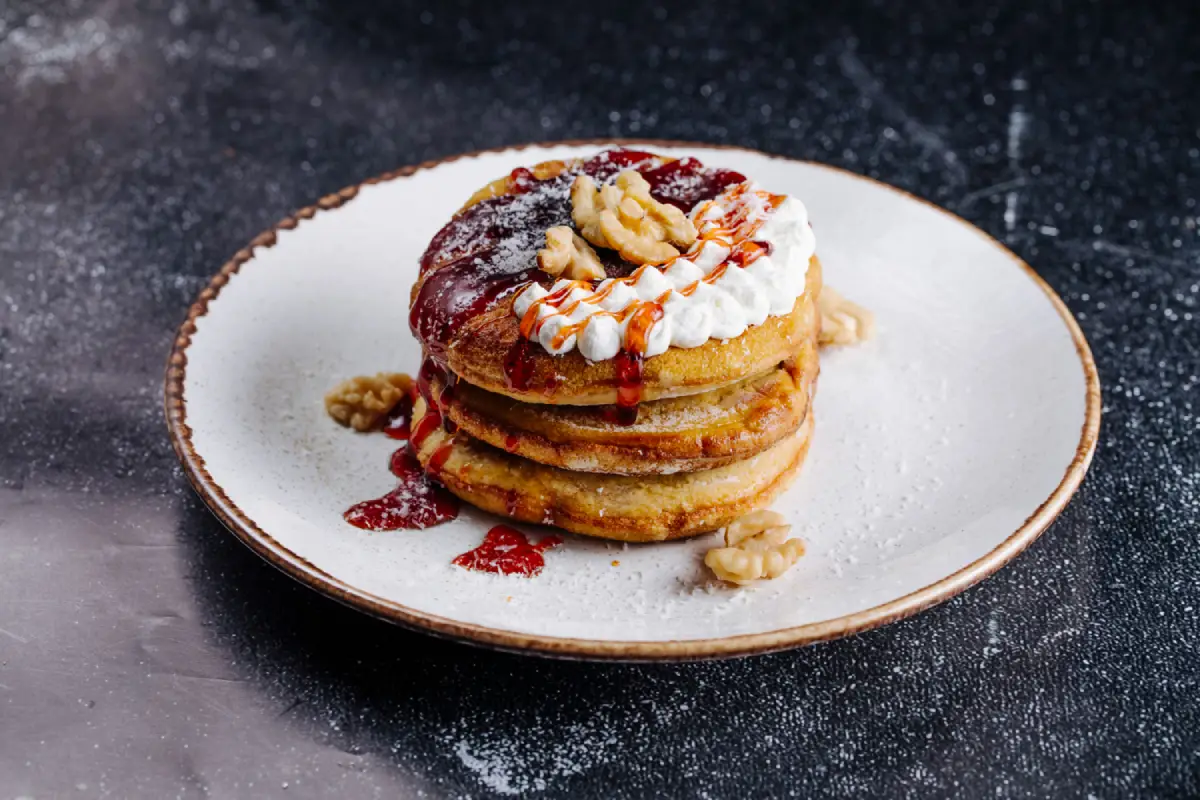A stack of mini pancakes adorned with vanilla cream, strawberry sauce, and walnuts on a speckled ceramic plate.