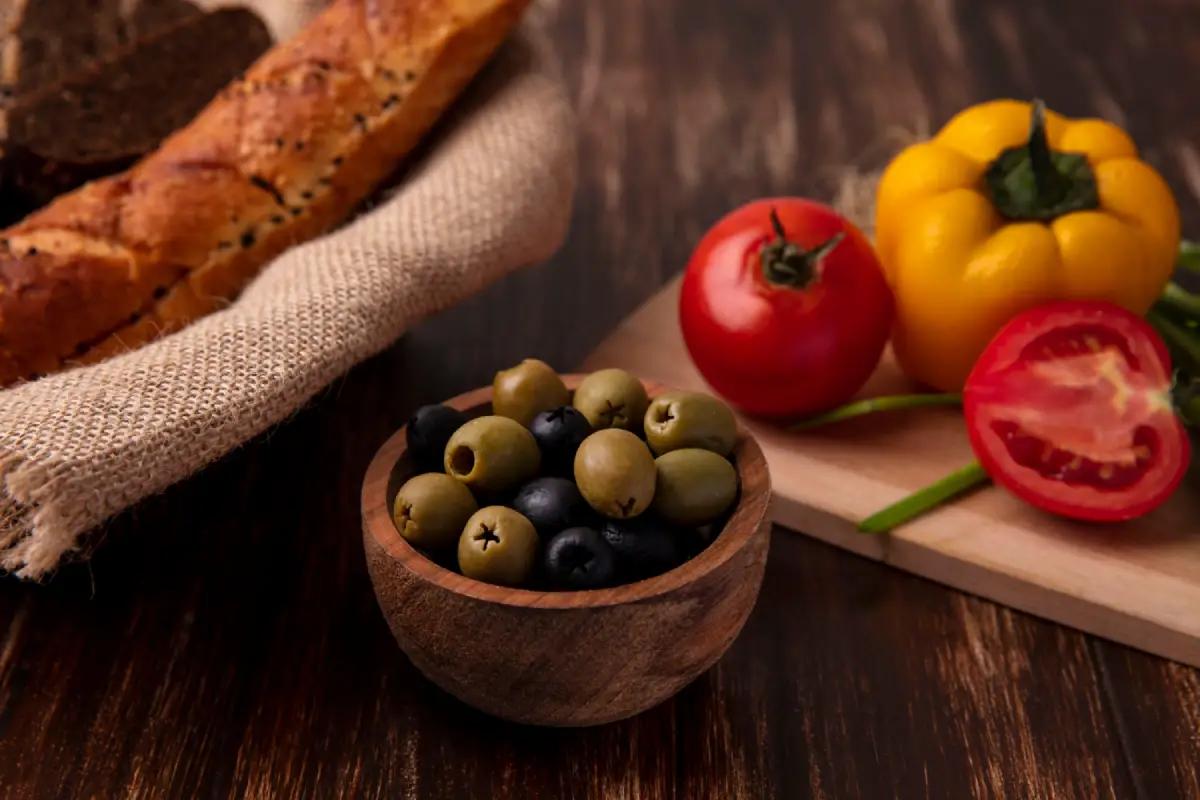A bowl of green and black olives with tomatoes and yellow bell pepper on a wooden table.