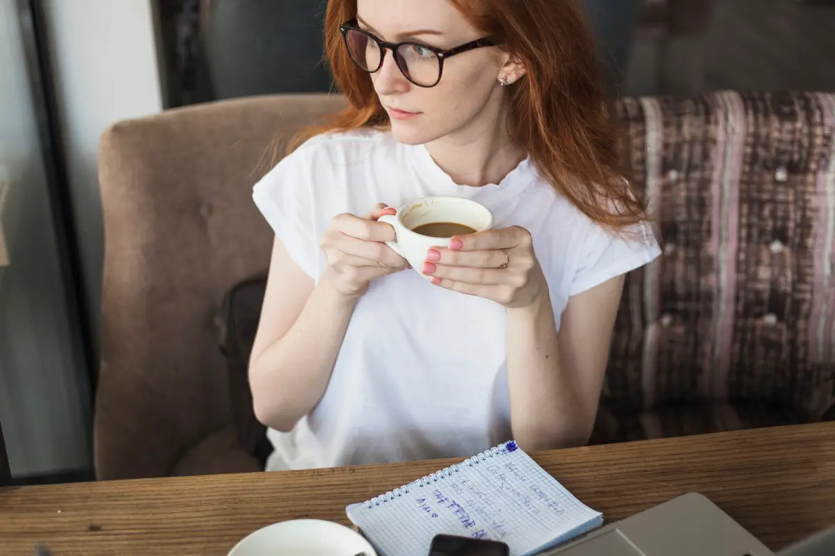 Red-haired woman thoughtfully sipping tea while reading notes.