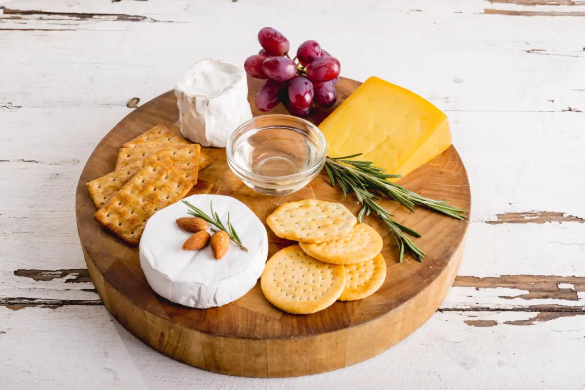A high-angle view of a cheese platter with grapes, herbs, and crackers on a wooden board.