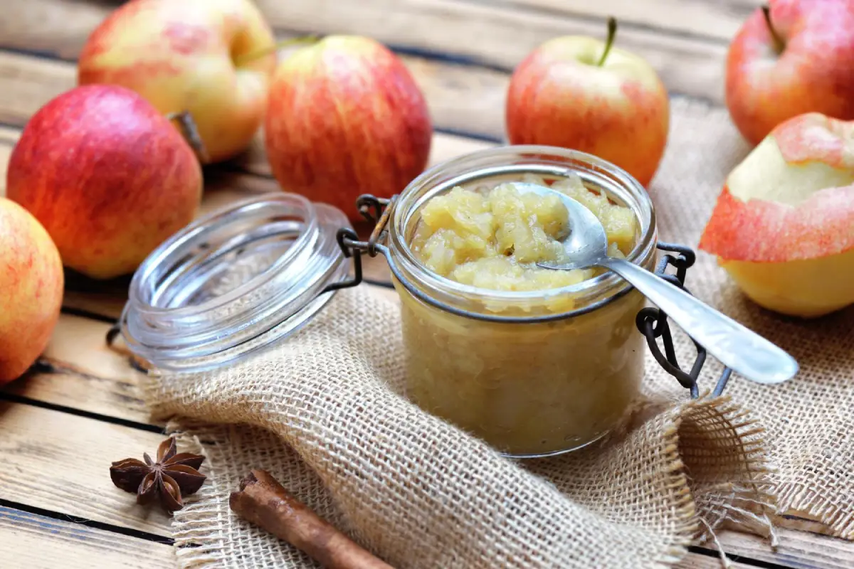 Homemade apple sauce in a glass jar with a spoon, surrounded by fresh apples, cinnamon sticks, and star anise on a rustic wooden table.