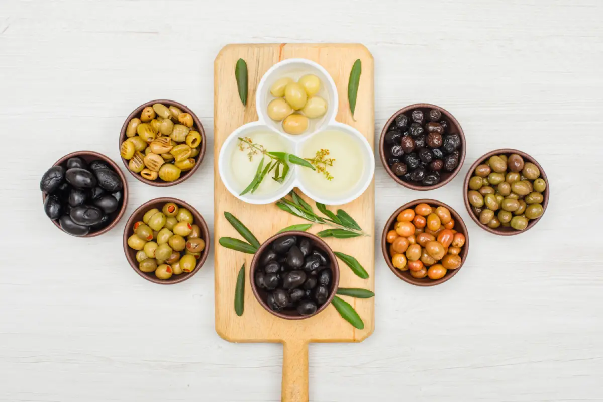 Assorted marinated olives and olive oil on a cutting board, surrounded by olive leaves.