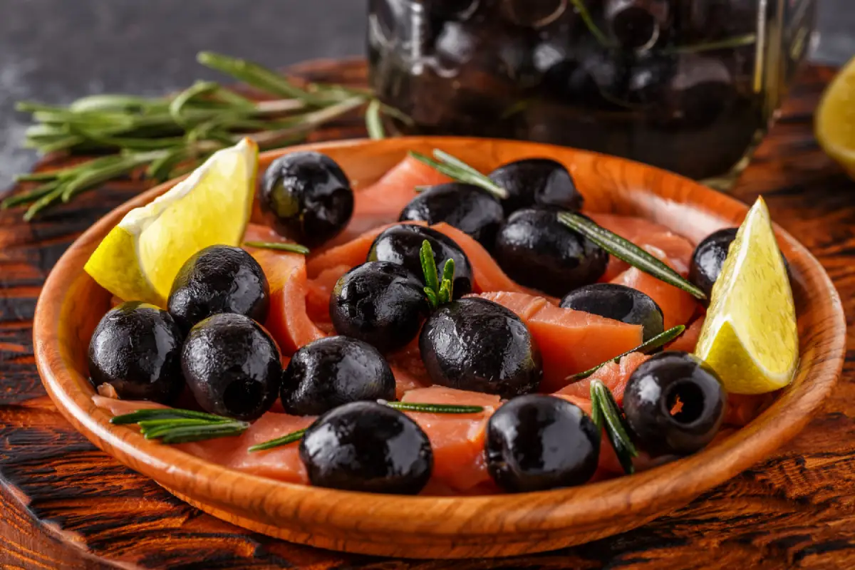 Fresh salmon slices with black olives and lemon wedges on a wooden plate.