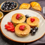 Mini pancakes adorned with fresh strawberries and a red heart-shaped berry on a grey plate, accompanied by a bowl of blueberries and honey.