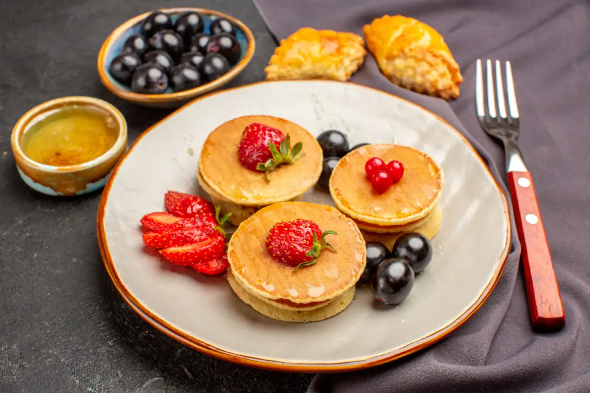 Mini pancakes adorned with fresh strawberries and a red heart-shaped berry on a grey plate, accompanied by a bowl of blueberries and honey.